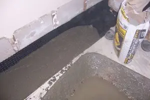 Concrete is being poured along a basement wall with exposed insulation, next to a bag of cement mix. The setting feels industrial and unfinished.