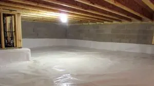 A clean, empty basement with exposed wooden beams and concrete block walls. The floor is covered with a white vapor barrier, and a single light bulb illuminates the space.