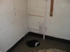 Basement corner with exposed concrete walls, a sump pump pit, and a white PVC pipe. The area appears utilitarian and slightly worn.
