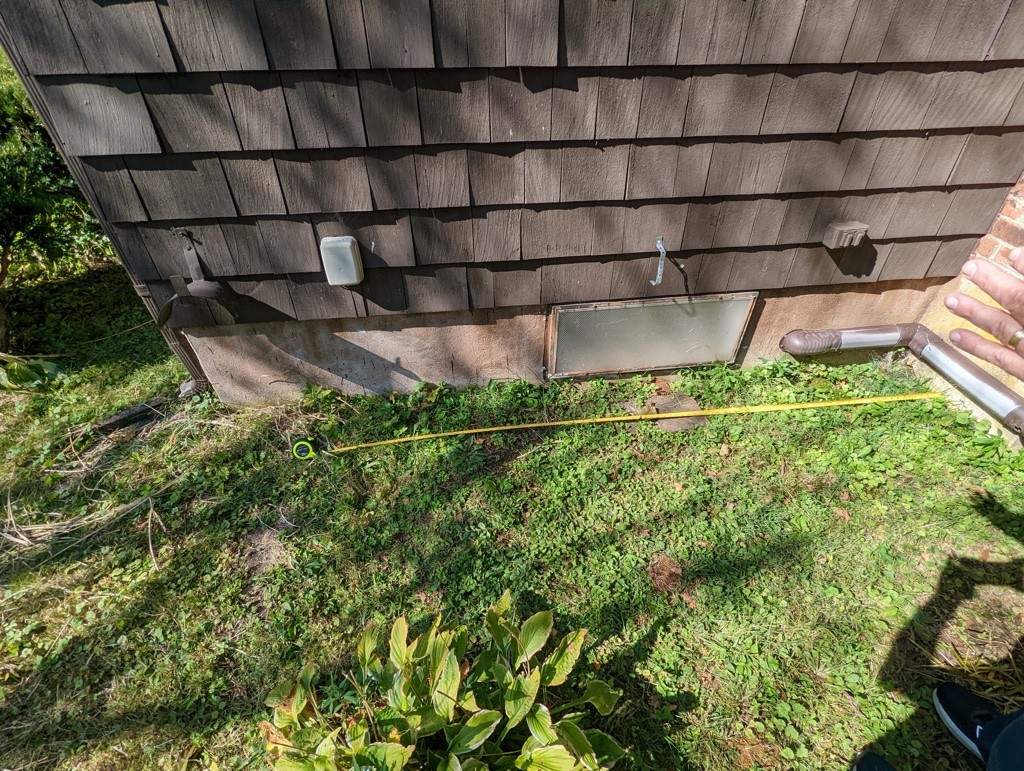 Side of a house with brown shingles, a vent, and greenery on the ground. A yellow tape measure extends across the grass, capturing a sunny, outdoor setting.