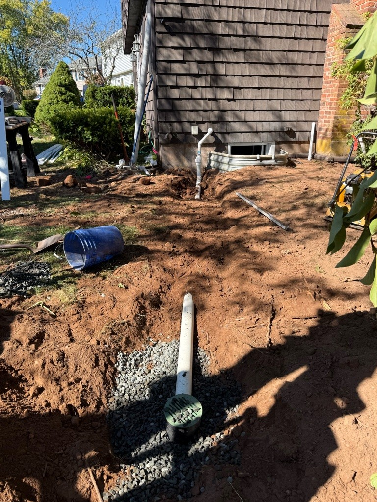 A backyard is under construction with dug-up soil and gravel. Pipes are being installed near a house's shingled wall and a blue bucket is nearby. Bright, clear day.