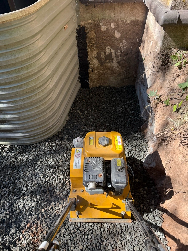 A yellow plate compactor sits on loose gravel in a corner with an earth wall and a light corrugated structure. Sunlight casts shadows on the ground.
