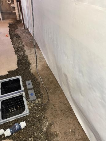 A basement waterproofing system is shown with drainage channels, gravel, and a white vapor barrier on the wall, conveying a sense of renovation and protection.