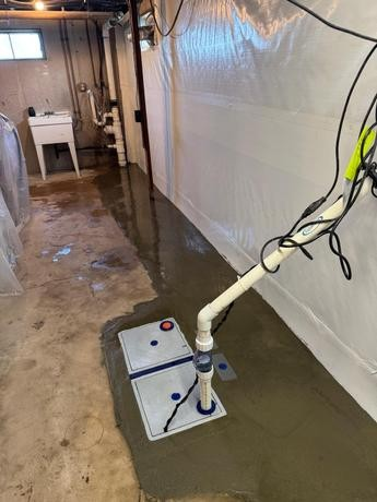 Basement with wet concrete floor and sump pump installation. Plastic sheeting lines walls; a utility sink is visible, conveying a damp atmosphere.