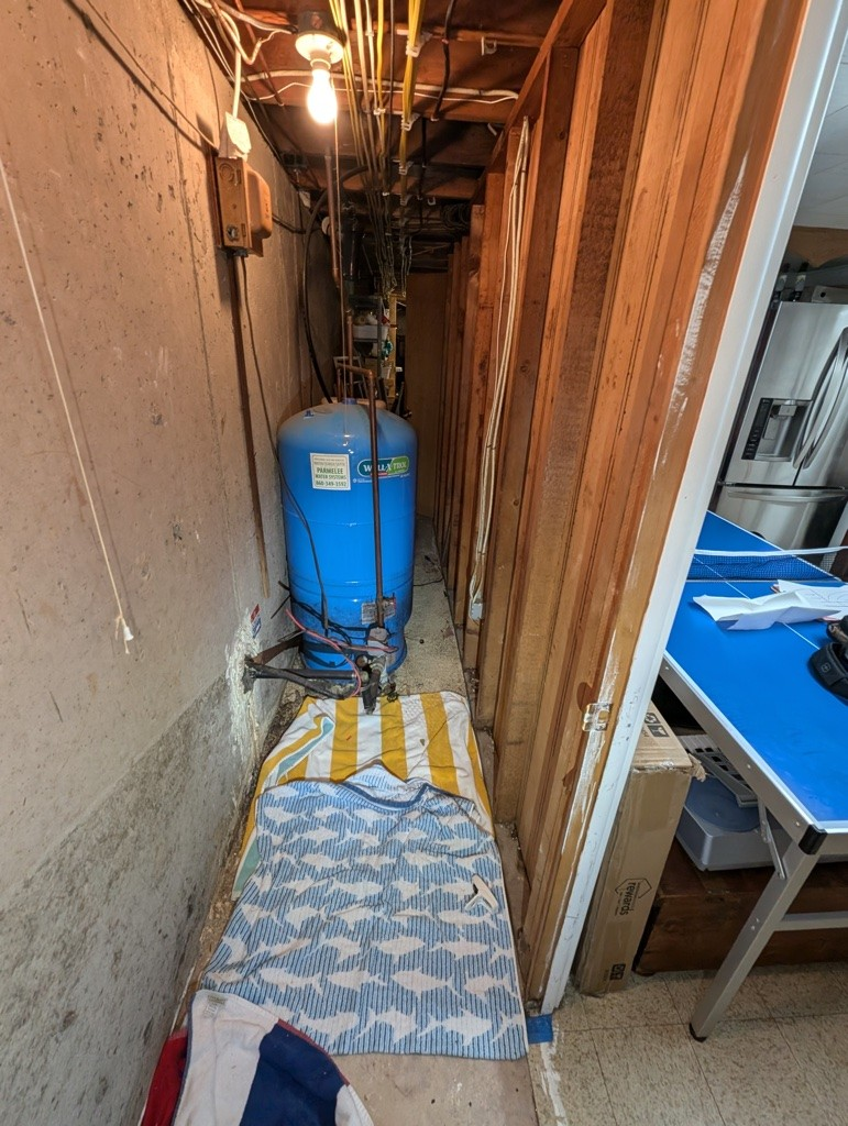 Narrow basement space with exposed pipes and a blue water tank. A lit bulb dangles overhead. Towels and a doorway lead to a room with a table.