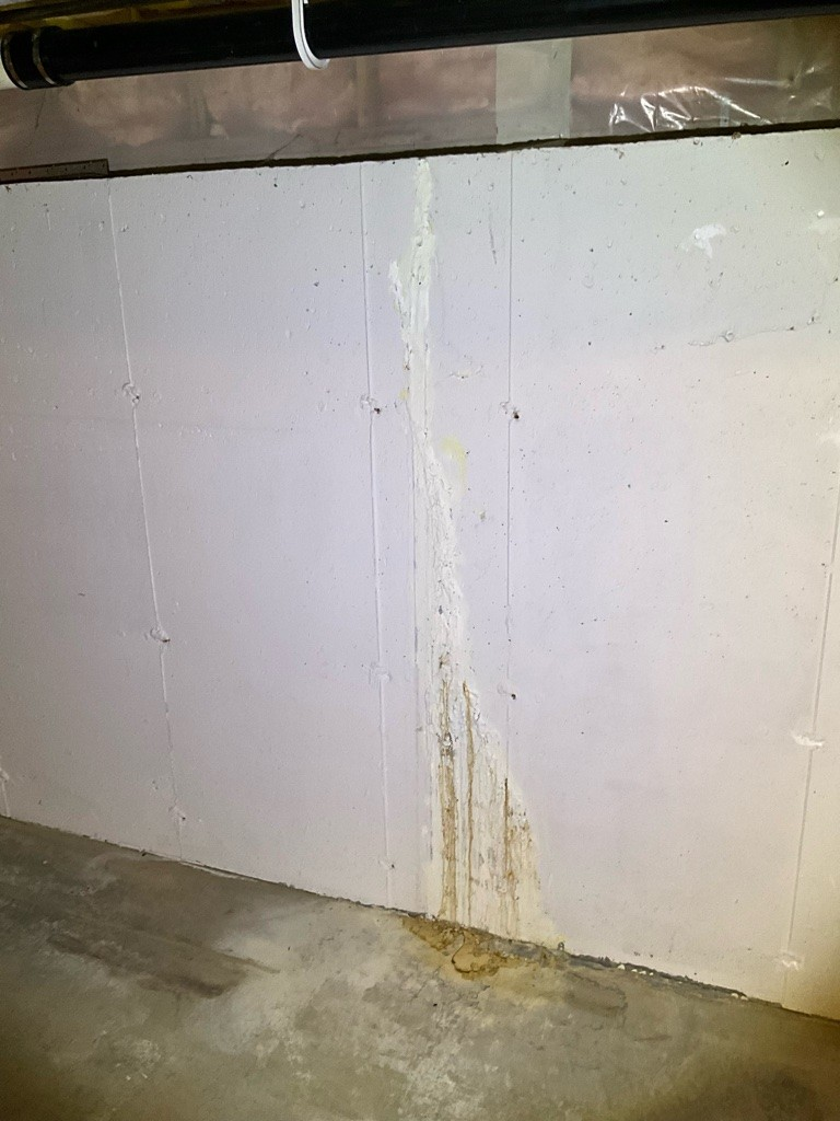 Concrete basement wall with vertical streaks of water damage, showing rust-colored stains. A black utility pipe runs horizontally near the ceiling.