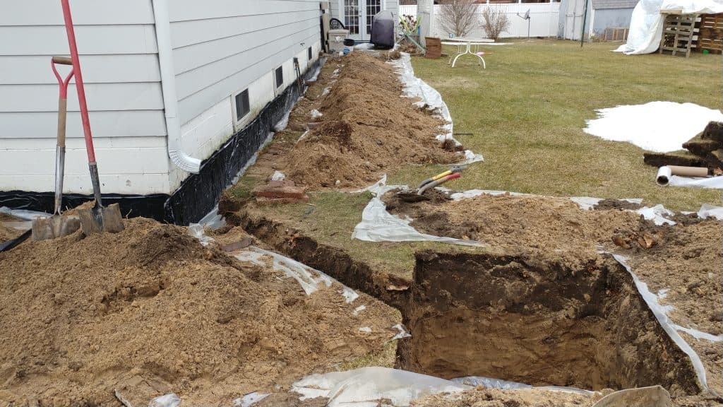Excavation site by a house with a long trench and pile of dirt. Shovels and plastic sheeting visible. Overcast day, with a grassy yard in the background.