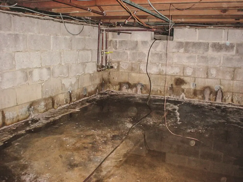 Flooded basement with concrete block walls and wooden beams overhead. Water covers the floor, indicating possible leaks or drainage issues.