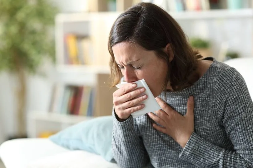 Woman holding a tissue to her face, appearing distressed, in a home setting with bookshelves, illustrating the impact of poor indoor air quality on health.