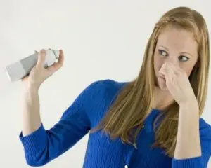 Woman in a blue sweater holding a spray can and pinching her nose, reacting to unpleasant odors, symbolizing indoor air quality issues.
