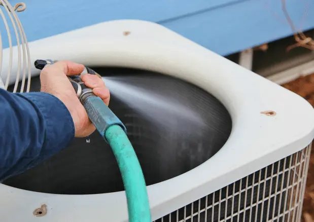 Person using a garden hose to clean the exterior of an air conditioning unit, emphasizing maintenance for optimal indoor air quality and ventilation.