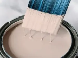 Paintbrush dipping into a can of light beige paint, illustrating the topic of low VOC and no VOC paint options related to indoor air quality.