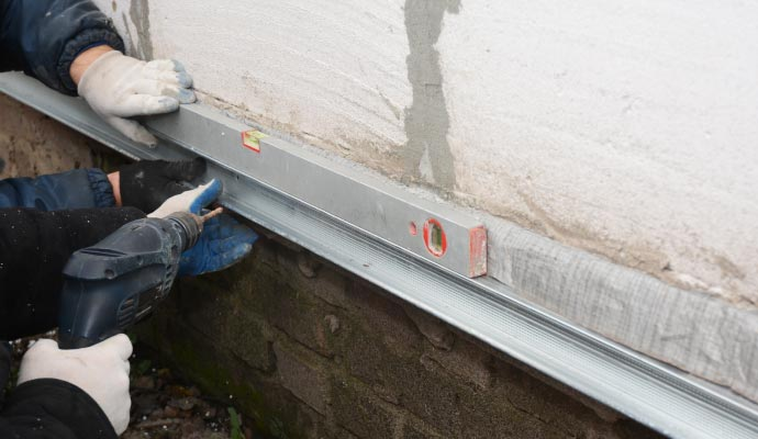 Workers installing a foundation repair system using a level and drill, demonstrating professional foundation reinforcement techniques for structural stability.