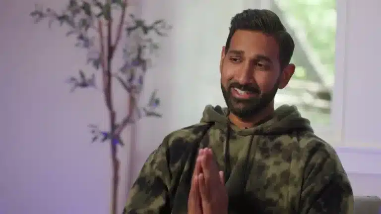Smiling person in hoodie with hands clasped.