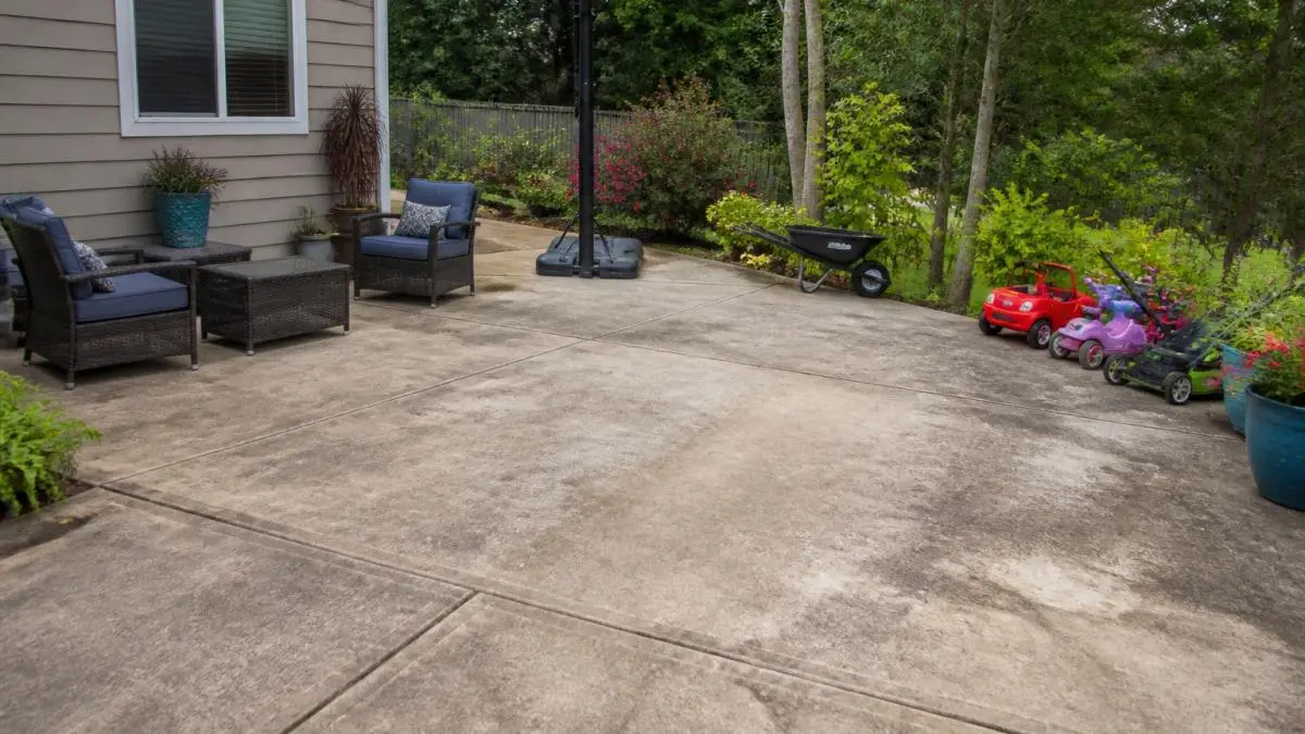 Residential driveway and patio area with scattered lawn furniture, colorful potted plants, children's toys, and garden equipment positioned around the perimeter before property preparation