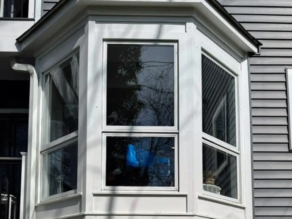 Image of a residential home's exterior featuring a modern bay window installation with white framing, surrounded by gray siding and decorative stones at the base.