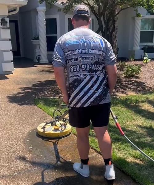 Man pressure washing a home's walkway with a surface cleaner. He wears a "Florida Pro Wash" shirt, advertising residential and commercial exterior cleaning services. 850-919-WASH.