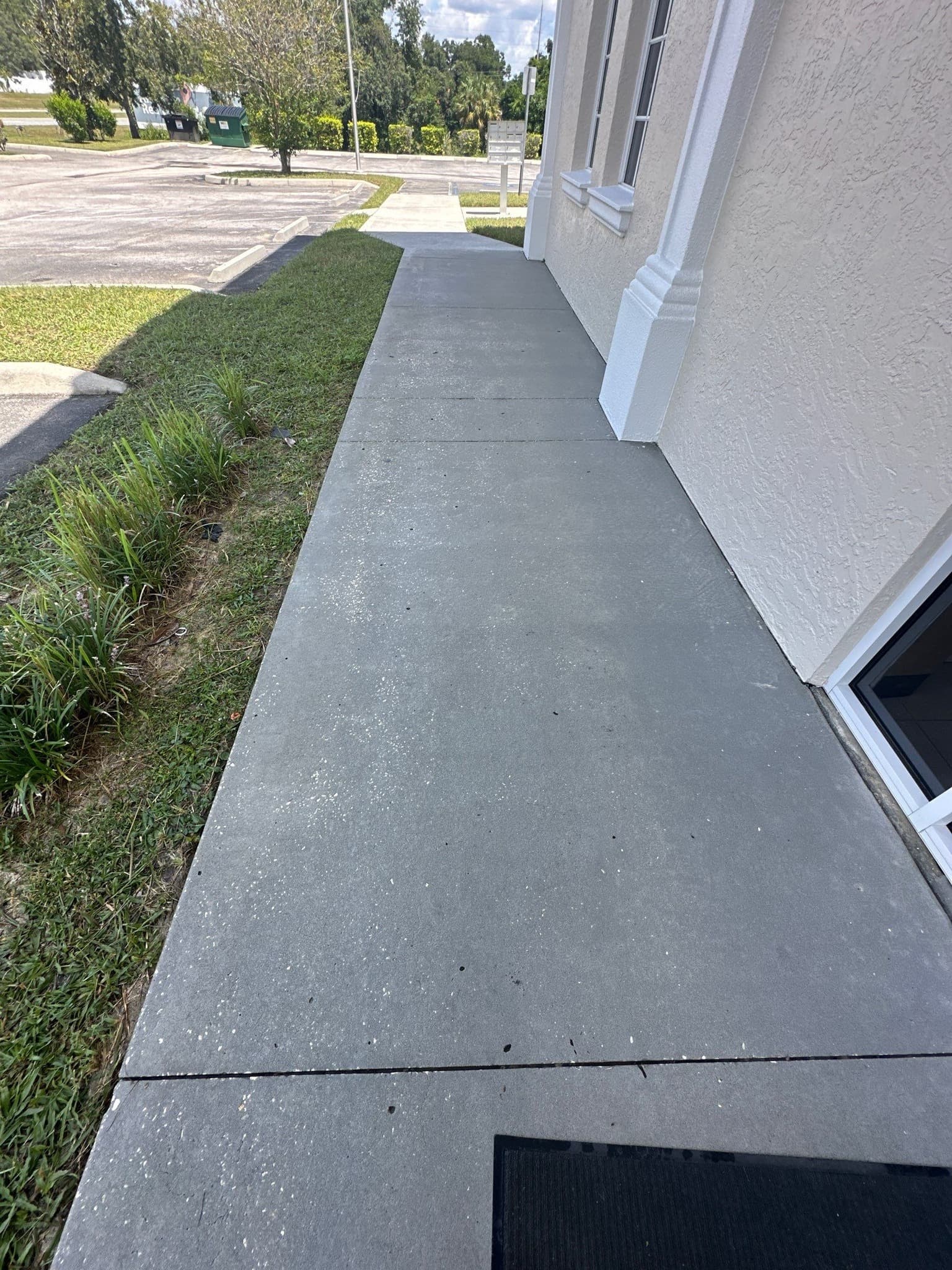 Clean, pressure washed sidewalk in Miramar Beach, FL, showcasing the results of pressure washing Miramar Beach FL services. Gray concrete runs alongside green grass and a building with white trim.
