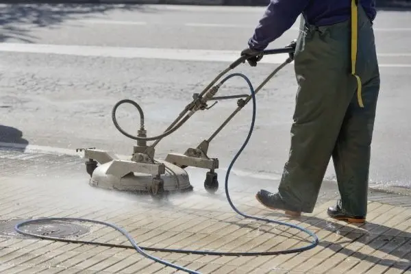 Man power washing a tiled sidewalk with a surface cleaner. The tool has a rotating spray bar under a circular housing. He's wearing waterproof overalls and boots.