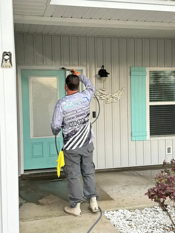 A technician from a Panama City FL exterior cleaning service soft washes a light blue front door. The company's logo is visible on his shirt: "Florida Pro Wash, 850-919-WASH".