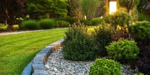 Illuminated Garden Landscape Design - Search Strategy Marketing - Search Strategy Marketing Manicured garden bed with stone edging, decorative gravel, lush green shrubs, manicured lawn, and warm outdoor lighting at dusk.