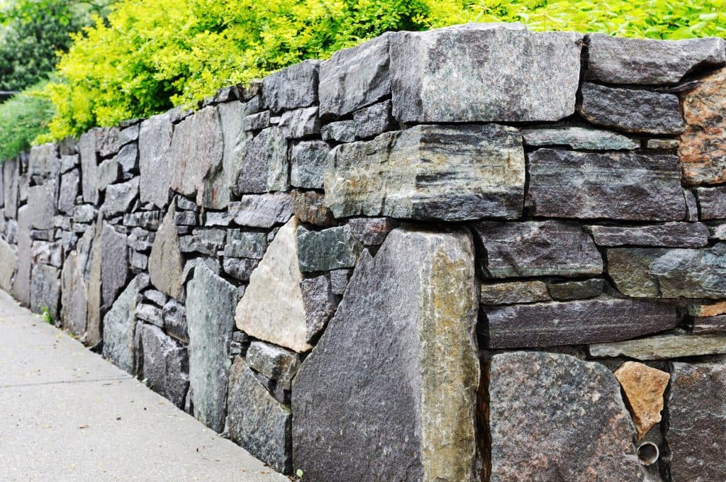 A close-up view of a custom-built stone retaining wall next to a sidewalk with green foliage along the top.