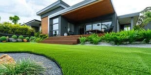 Modern Home Landscaping Design - Search Strategy Marketing Modern house with a wooden deck extending to an outdoor seating area, surrounded by lush green lawn and tropical plants.
