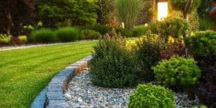 Illuminated Garden Landscape Design - Search Strategy Marketing Manicured garden bed with stone edging, decorative gravel, lush green shrubs, manicured lawn, and warm outdoor lighting at dusk.