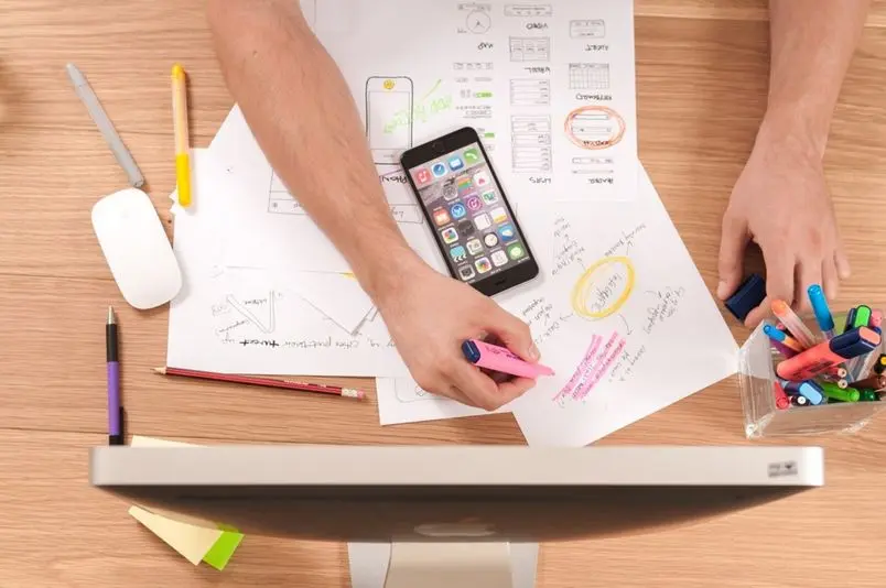 a man holding a pink highlighter with multiple sheets of paper and an iphone doing research