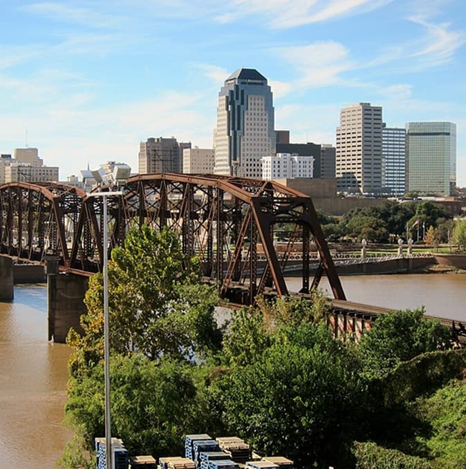 A steel truss bridge crosses a river with a city skyline featuring tall buildings in the background under a blue sky.