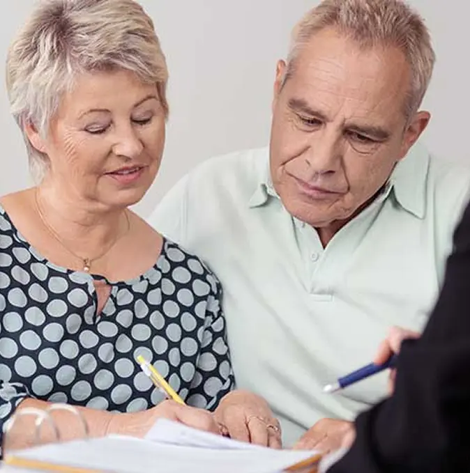 Two older adults sit together reviewing and signing documents while a third person, partially visible, holds a pen and explains the paperwork