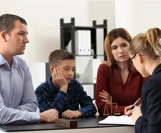 A family of three, looking serious, consults with a professional woman over paperwork in an office. A scale symbolizes justice on the desk.