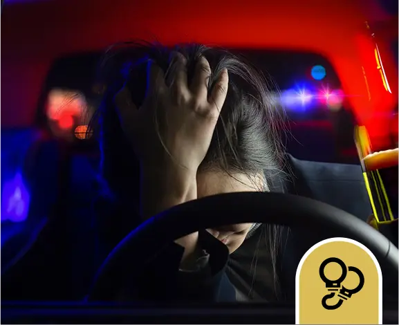 A person sits behind a car steering wheel with their head in their hand, police lights flashing in the background, and a drink with a straw visible in the corner.