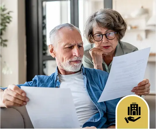 Elderly couple sitting on a sofa, both intently reading documents. The man looks concerned, while the woman, wearing glasses, appears thoughtful.