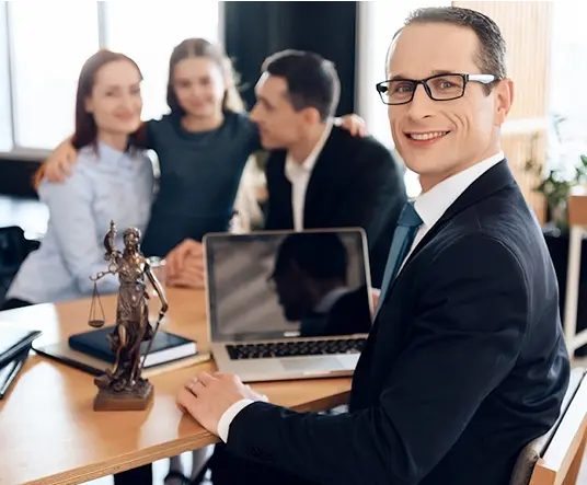 Smiling man in a suit sits at a desk with a laptop and statue, while a family in the background shares a warm moment, conveying trust and professionalism.
