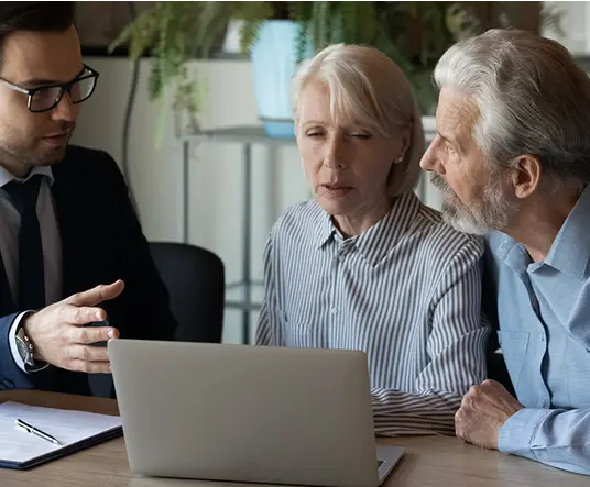 A young man in a suit discusses something on a laptop with a senior couple. The setting is an office with plants, conveying a serious and focused mood.