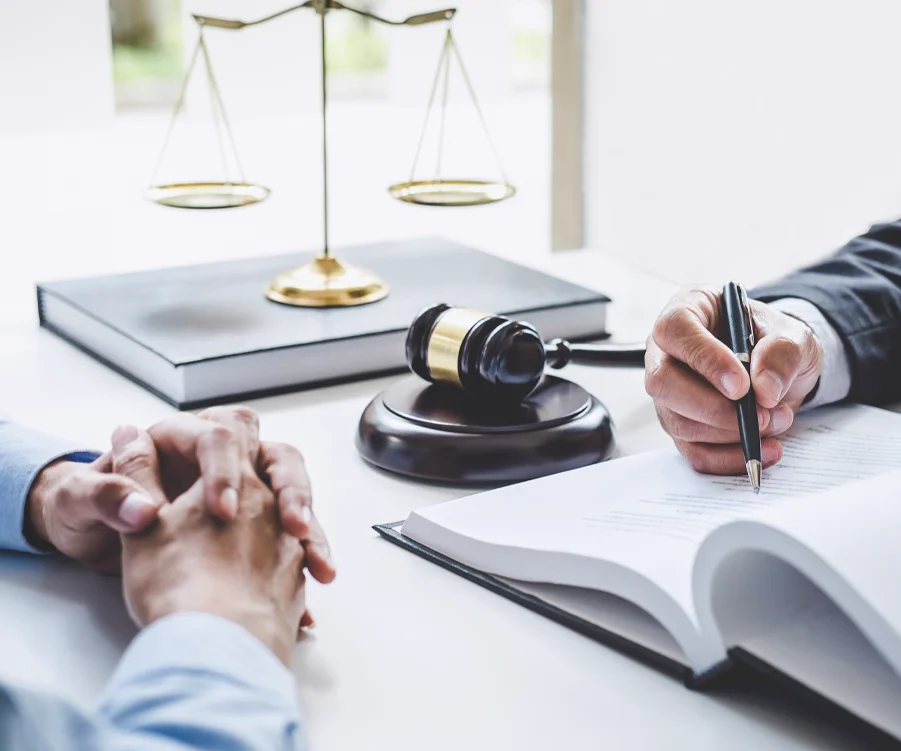two people sit at a desk with legal documents, a gavel, and scales of justice, suggesting a legal consultation or discussion.