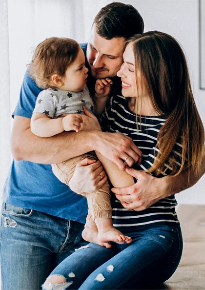 a family of three shares a warm moment; a child sits on an adult's lap while another adult hugs them both, smiling together.