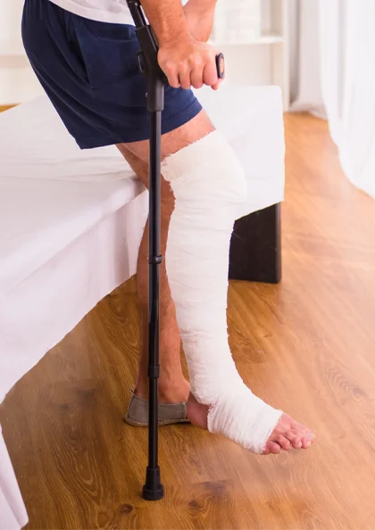 a man in shorts sits on a bed, holding a cane with a bandaged leg resting on the floor, indicating injury and support.