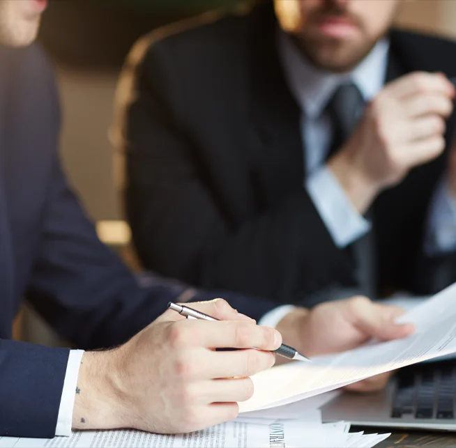 two business professionals discussing paperwork; one is writing notes while the other gestures, both are dressed in formal suits.