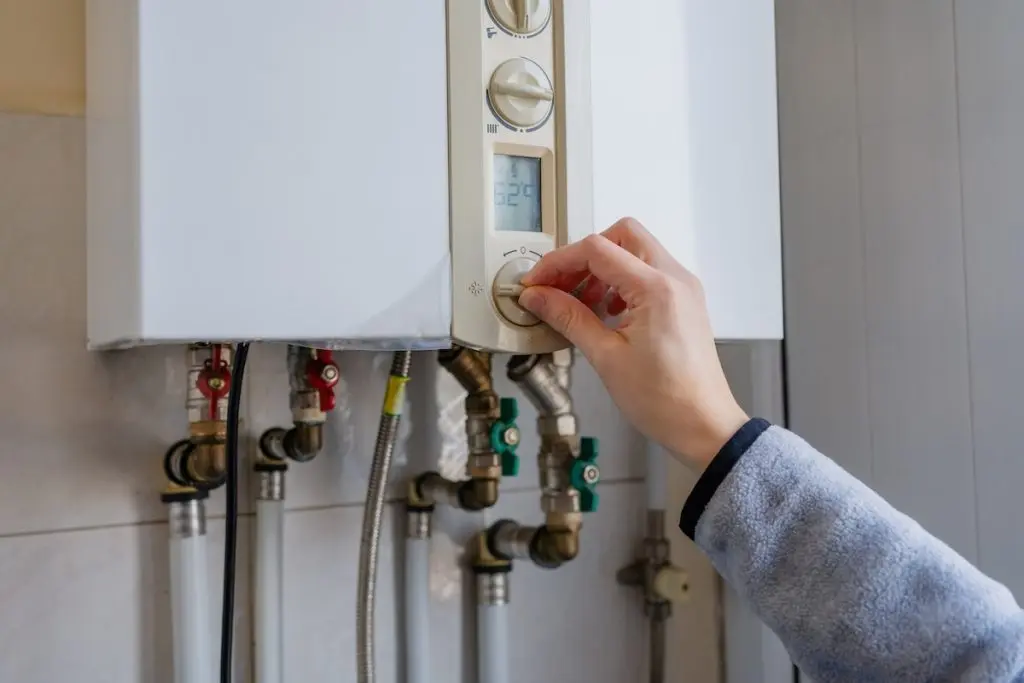 Hand turning the control knob on a white wall-mounted gas boiler, with pipes visible below, representing residential heating systems and everyday energy management at home.