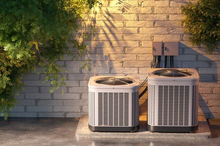 Two outdoor air conditioning units installed next to a brick wall with power connections and surrounding greenery. Scene relates to HVAC systems and home climate control.