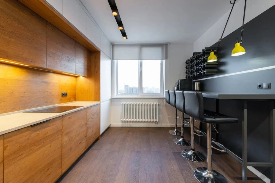 Contemporary kitchen interior with wooden cabinets, black bar stools, and large window.