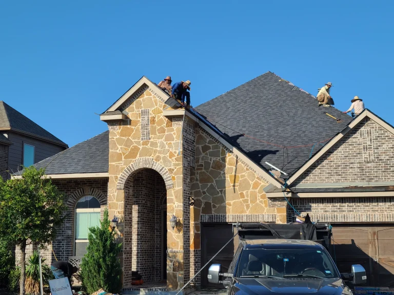 Multiple roofers installing dark gray shingles on a stone and brick home.