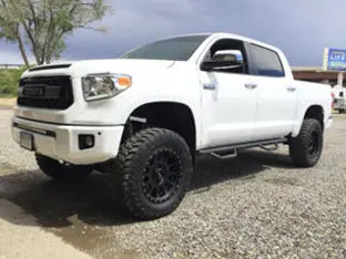 White lifted pickup truck parked on gravel with large off-road tires and black rims. Trees and a signboard in the background showcase one of our impressive customer rigs.