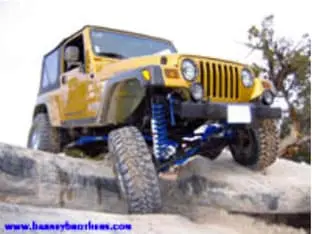 Yellow off-road Jeep navigating rocky terrain with suspension flexed, showcasing some impressive customer rigs, promotional website text in blue at the bottom left.