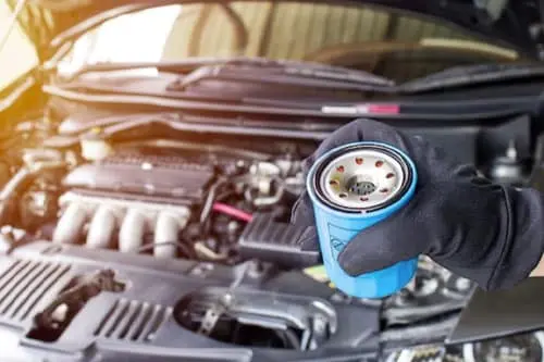 A gloved hand holds an oil filter in front of an open car hood with the engine visible.