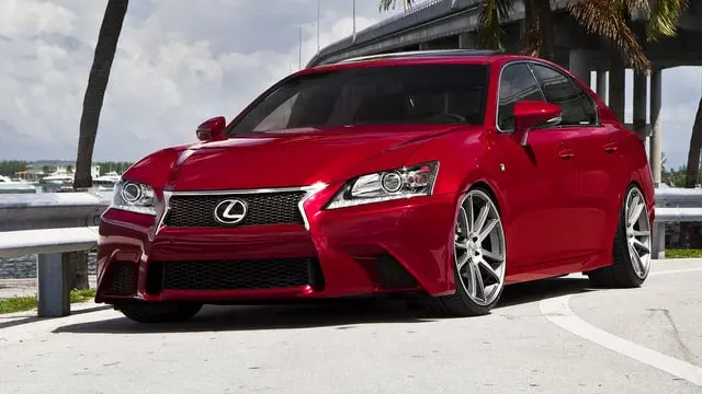 A red Lexus car parked near a waterfront with a bridge and swaying palm trees in the background.