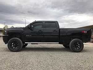 A black Chevrolet pickup truck with a crew cab and large off-road tires, parked on a gravel surface under a cloudy sky.