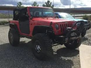 A lifted red Jeep Wrangler, one of the impressive customer rigs, with large off-road tires and a winch, is parked outdoors on gravel.